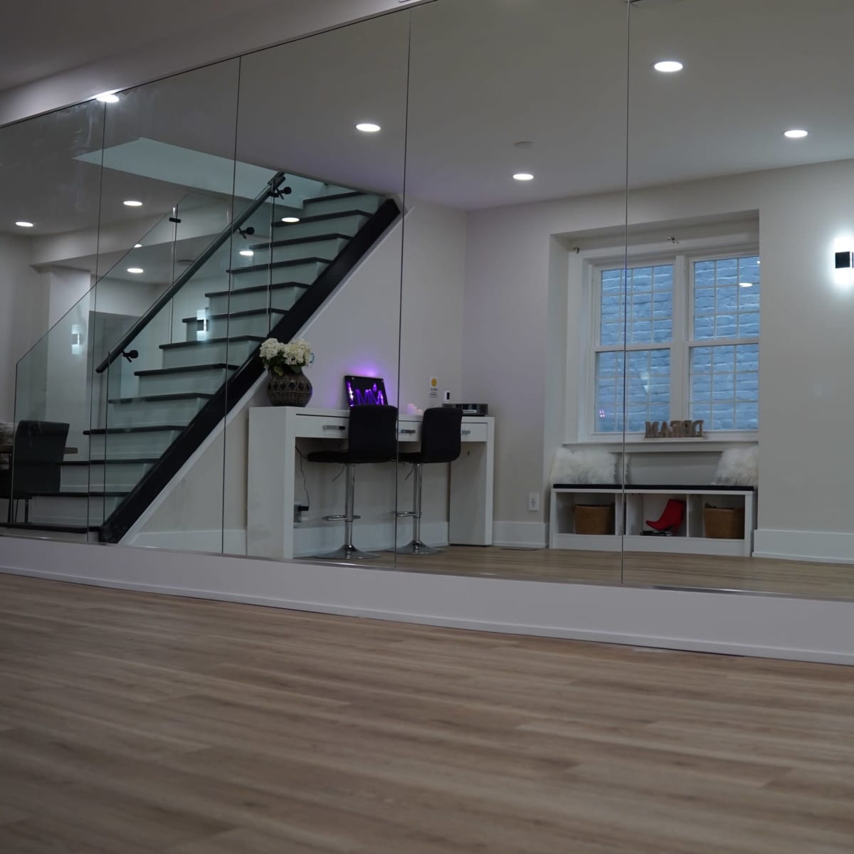 A modern room with wooden flooring, a large mirror on one wall, a staircase in the background, a workspace with two chairs, and a television mounted on another wall.