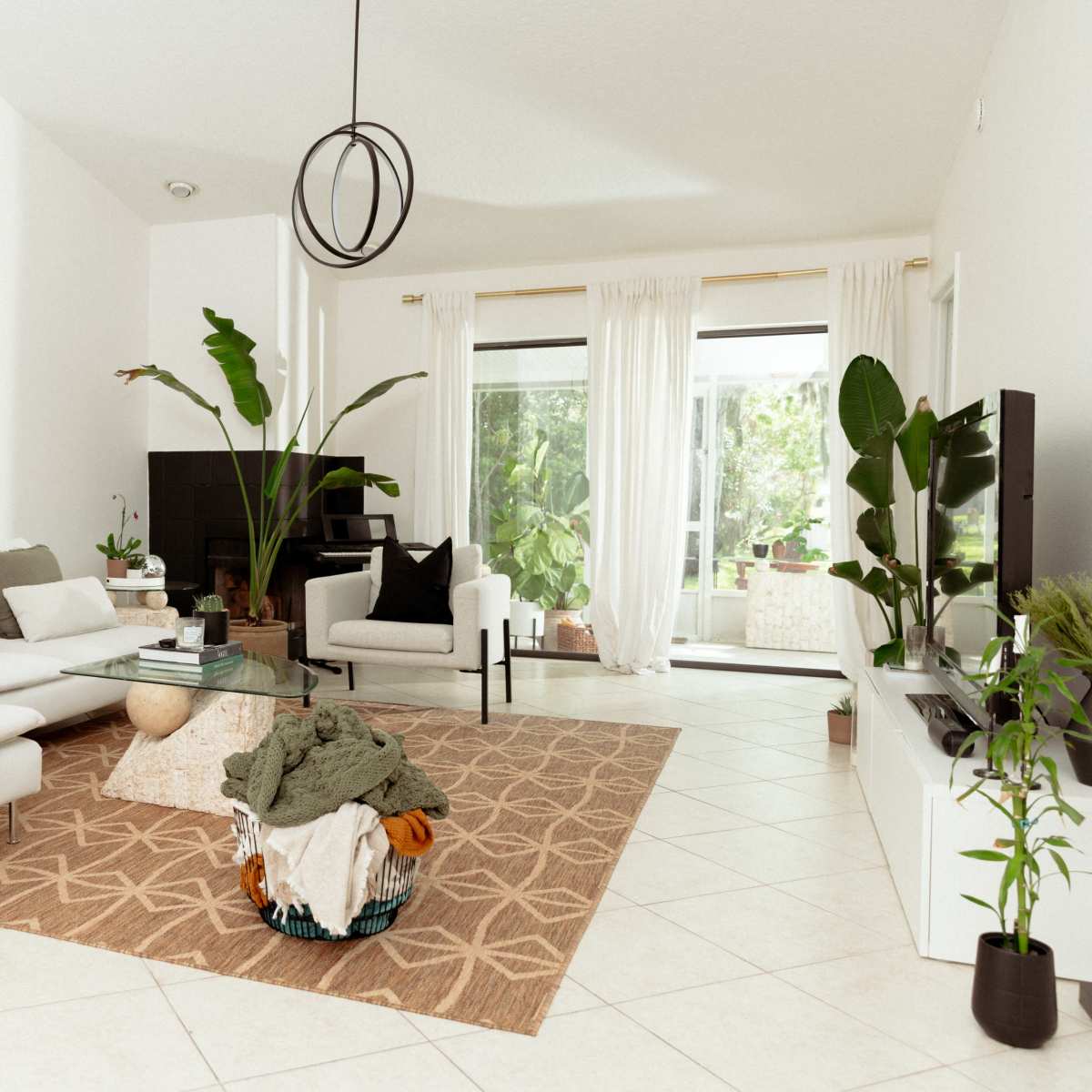 The living room features a white sectional sofa with a patterned rug, a coffee table, and large windows that let in natural light, surrounded by indoor plants.