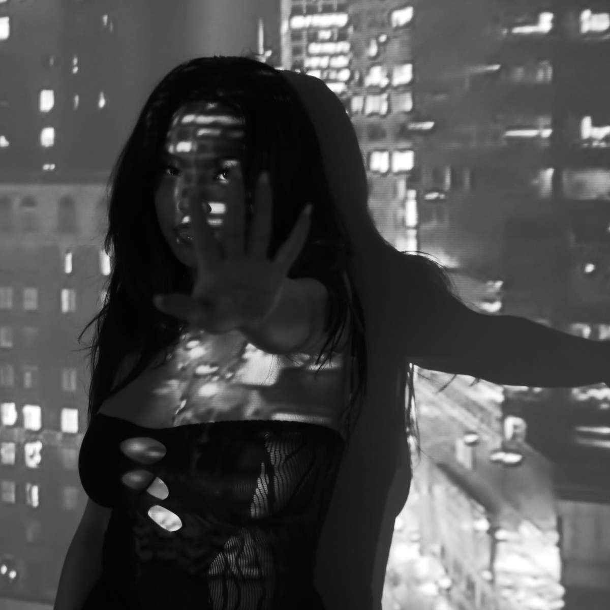 A woman stands in front of a cityscape projection, with her hand raised as shadows play across her figure.