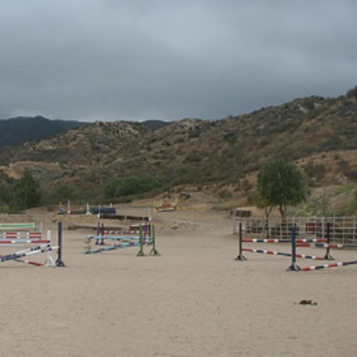 An equestrian arena with multiple colored jumps set up on sandy terrain surrounded by hills and overcast skies.