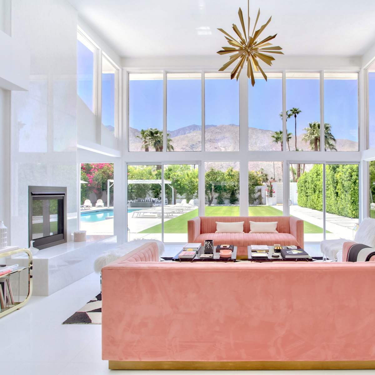 The living room features two large sofas facing a wall of windows that overlook a pool and palm trees with mountains in the background.