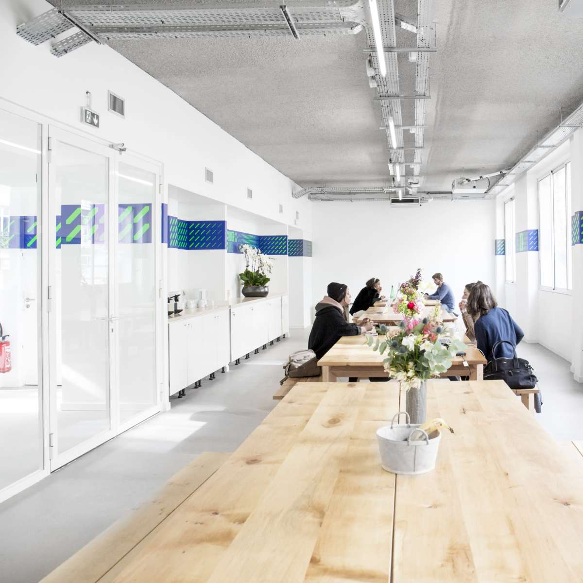 A long wooden table is surrounded by several people in a bright, modern office space with large windows and decorative wall designs.