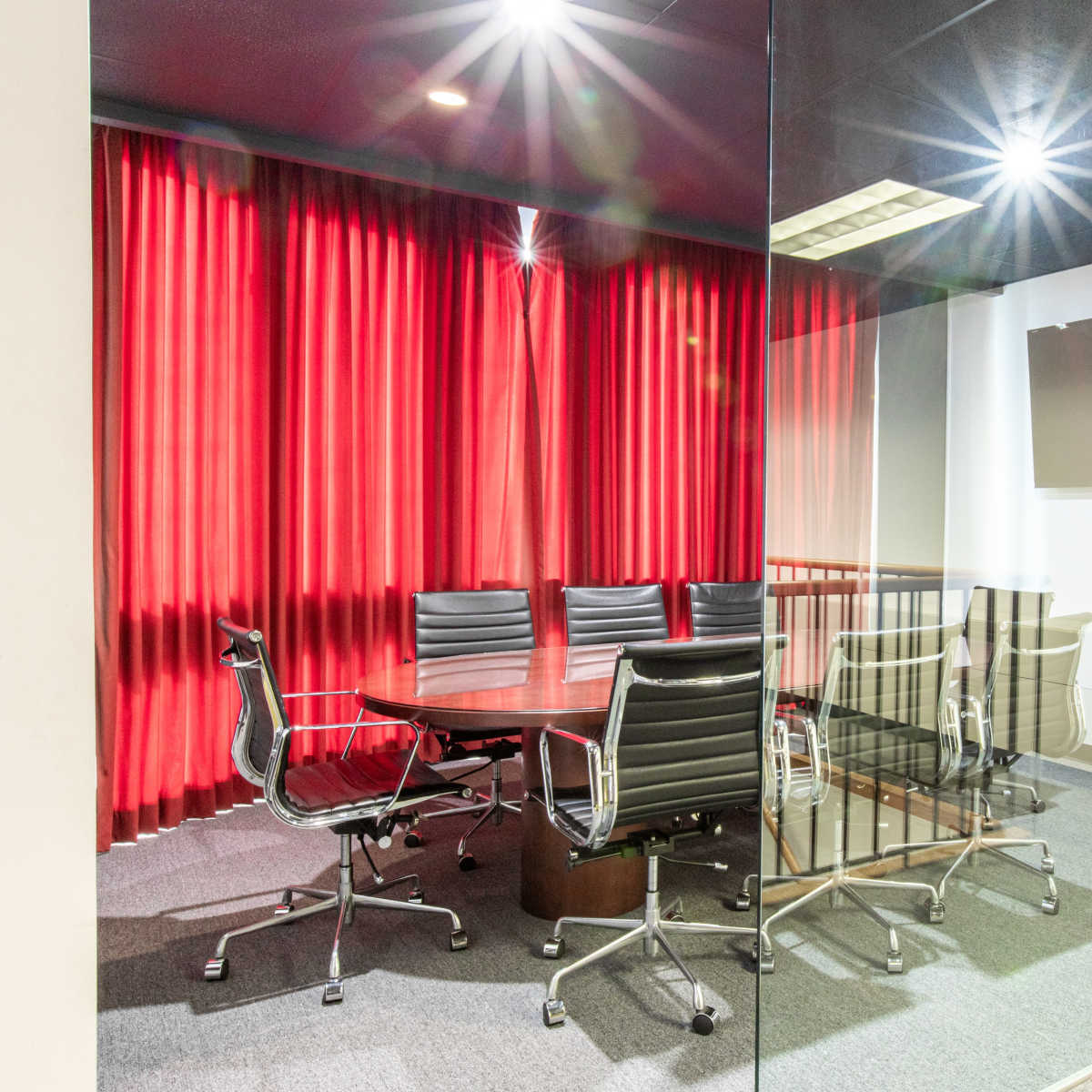 A glass-walled conference room features a large round table surrounded by black chairs and is illuminated by red curtains draping in the background.