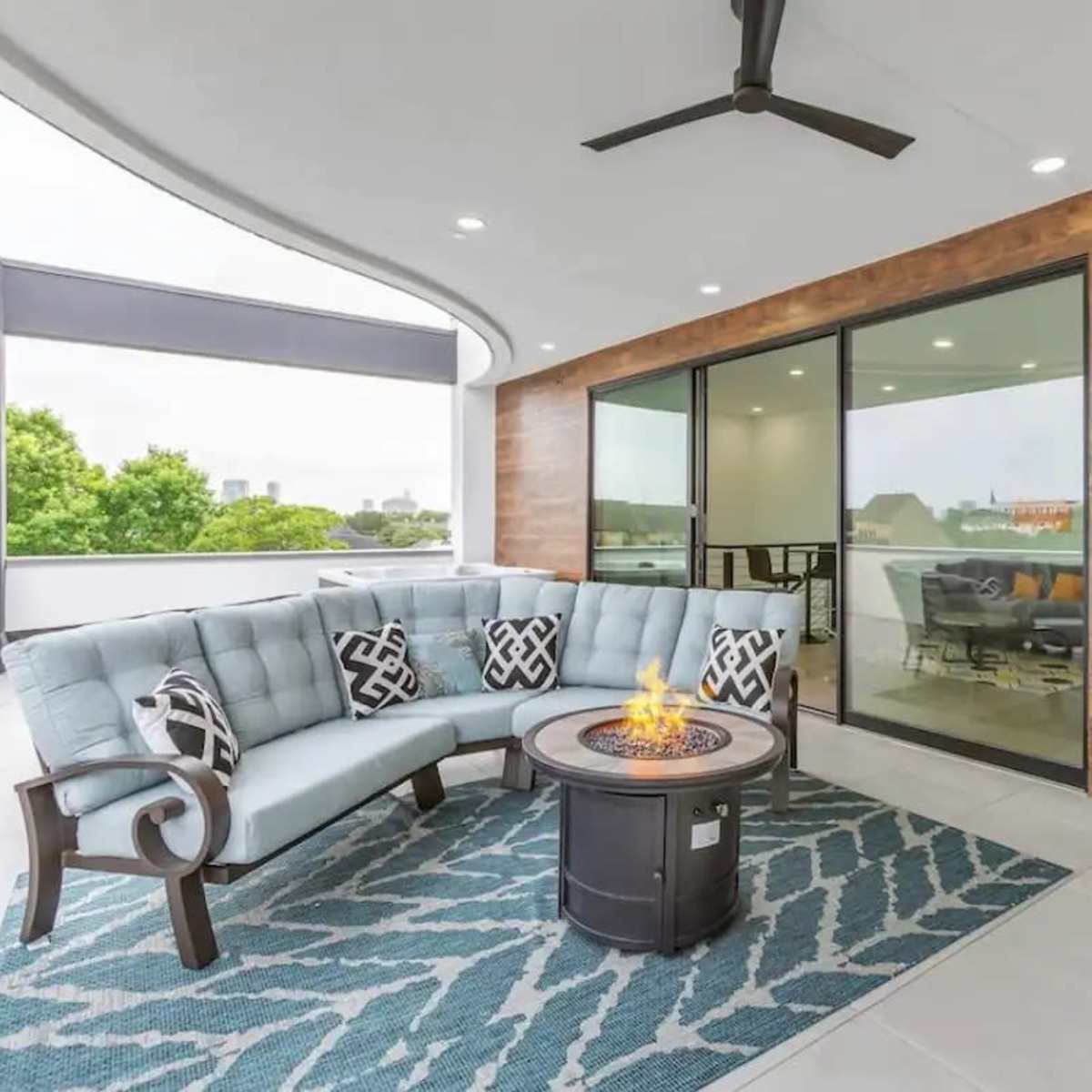 A modern balcony with a sectional sofa, a circular fire pit, and large glass doors leading into an interior space.