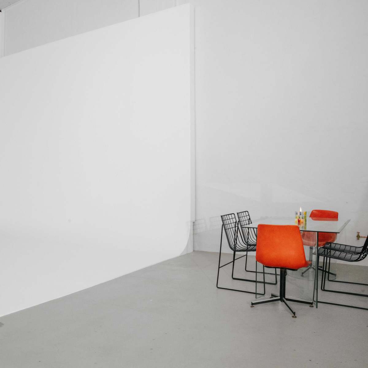 A minimalist studio space featuring a white backdrop, an orange sofa, a glass table with orange chairs, and a few drinks on the table.