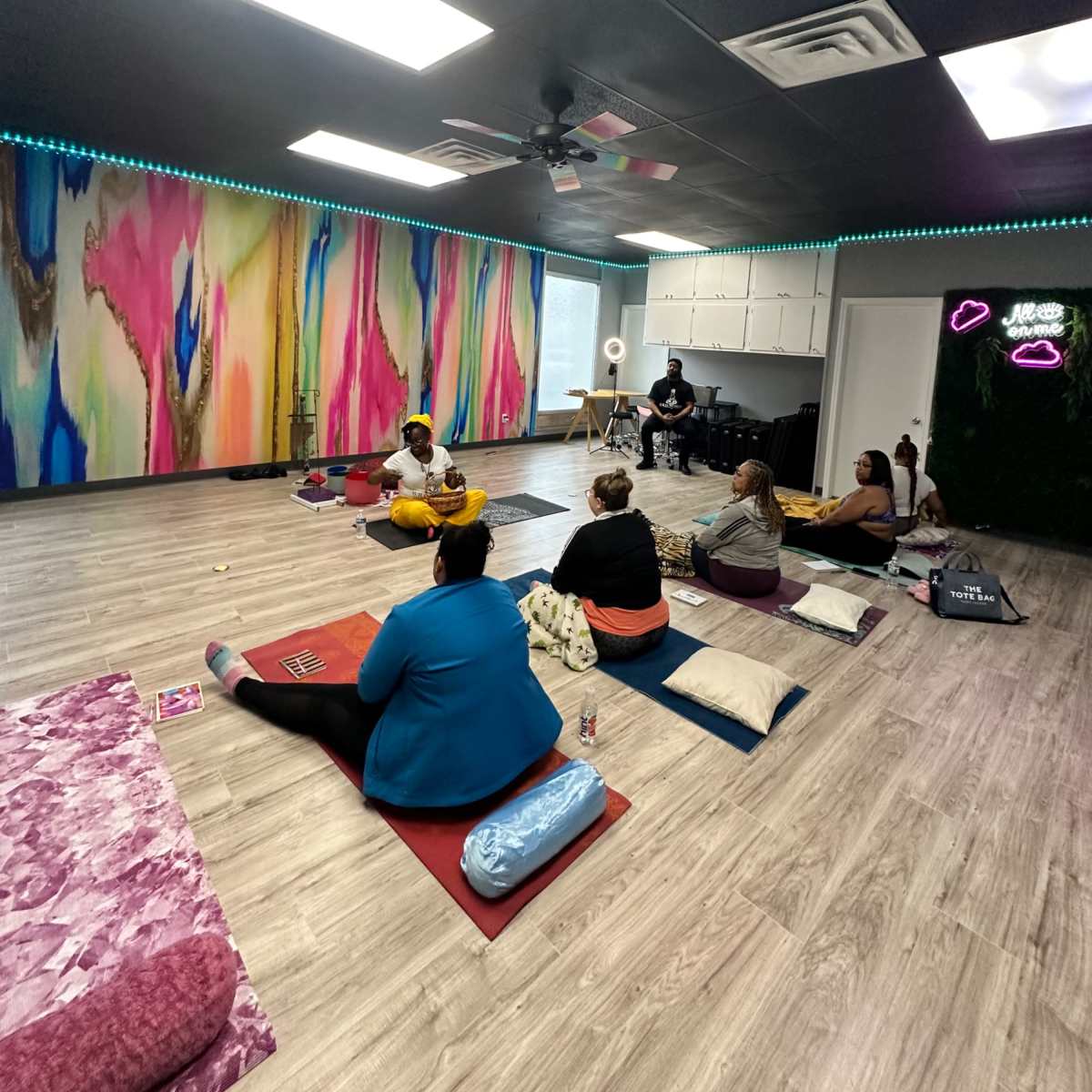 A group of people sits on the floor in a colorful, modern wellness studio, with vibrant wall art and yoga mats arranged around them.