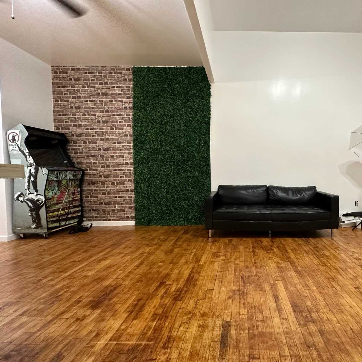 The scene features a spacious room with a black couch centered against a wooden floor, complemented by a faux brick wall and an artificial green wall on one side, along with two light stands nearby.