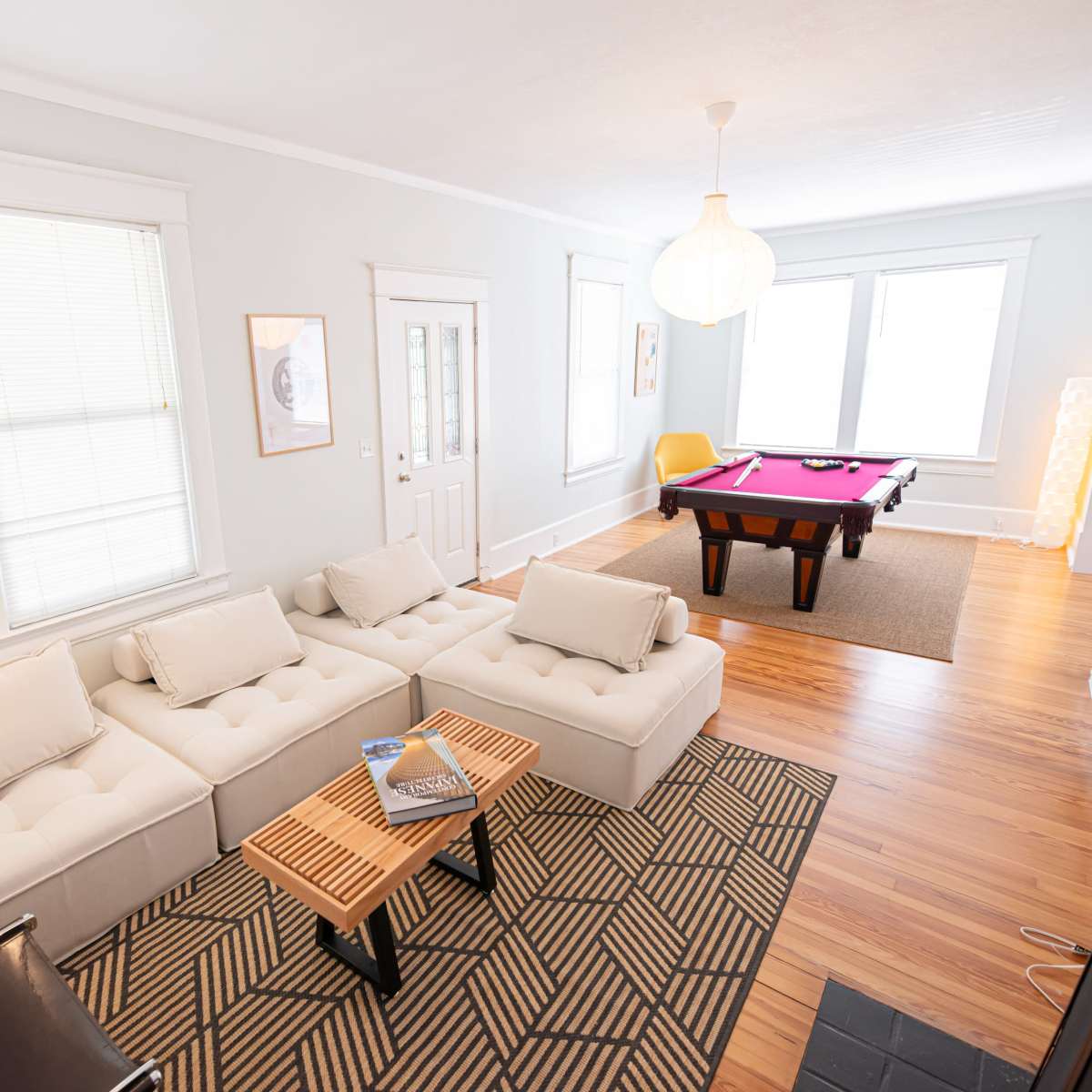 A well-lit living room featuring a light-colored sectional sofa, a pool table, and large windows with sheer curtains.
