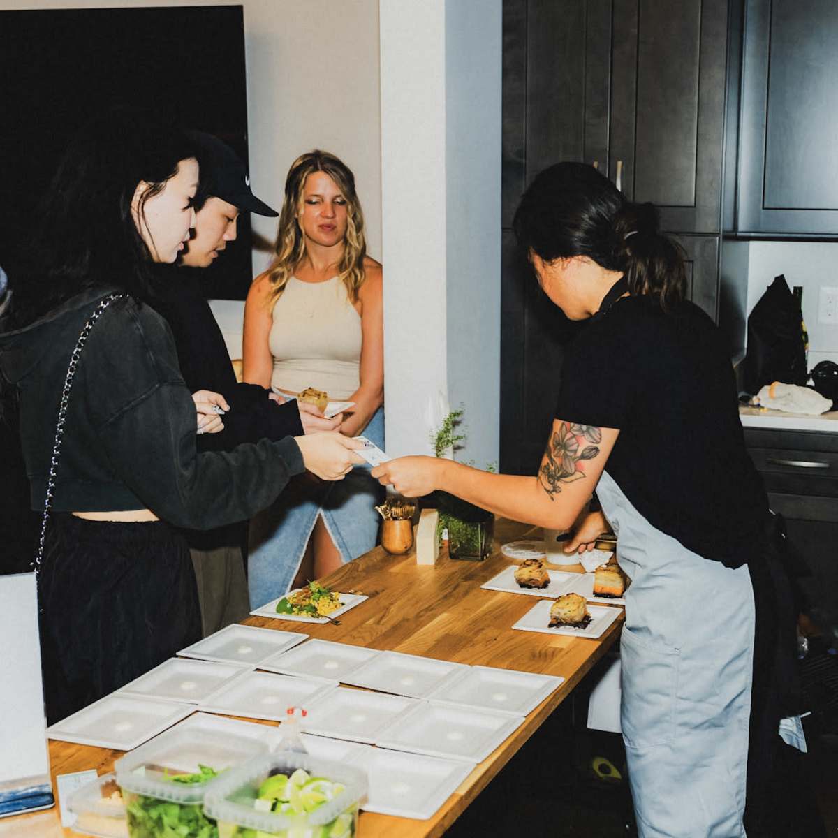 A woman hands a plate of food to another woman in a modern kitchen setting while additional guests are present in the background.