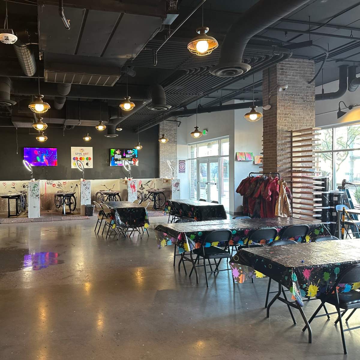 An indoor art studio with tables covered in floral tablecloths, exposed brick walls, and several illuminated screens displaying colorful images.