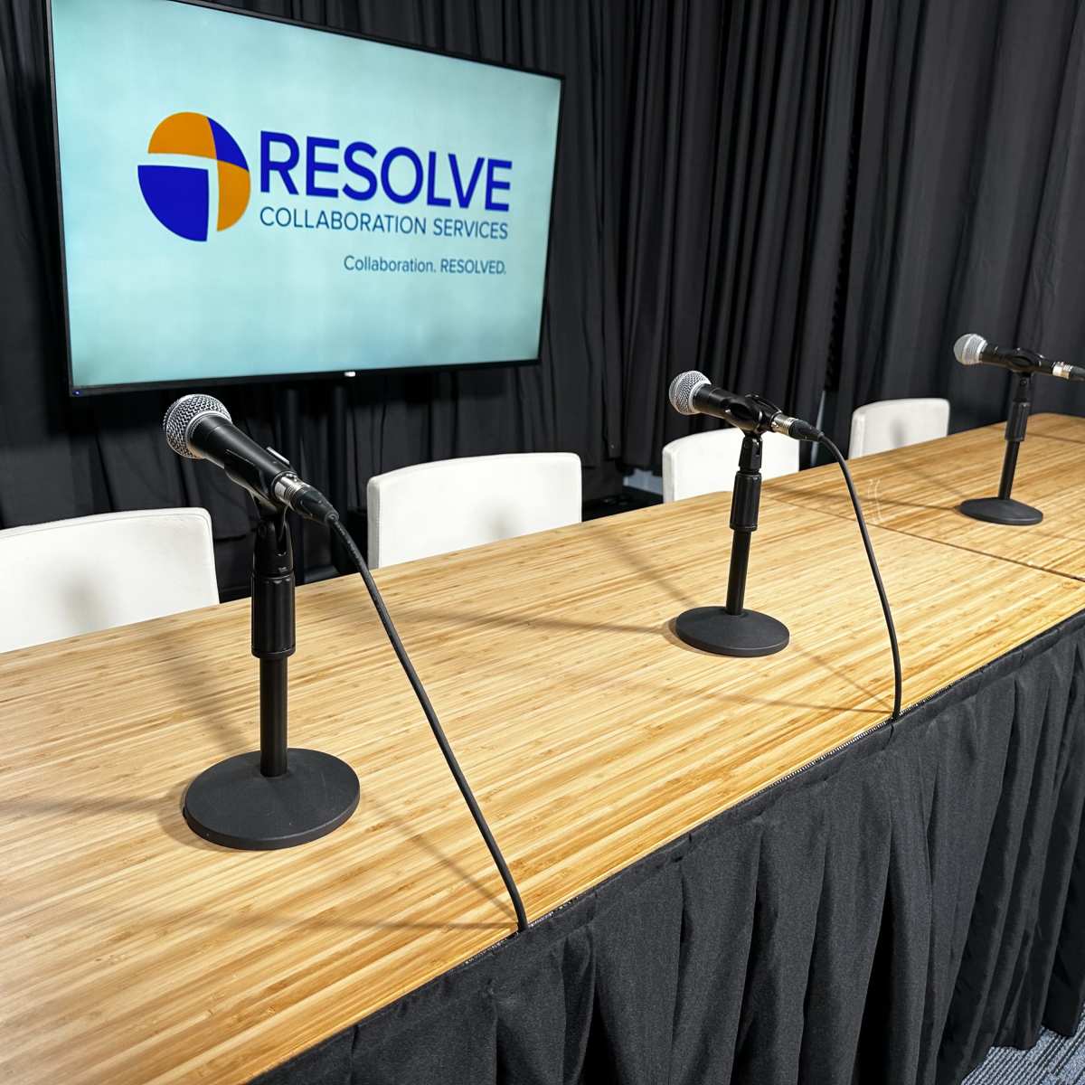 A wooden panel table with four microphone stands is set up in front of a screen displaying the logo of Resolve Collaboration Services.