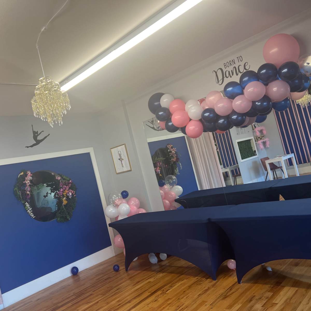 A decorated dance studio featuring blue walls, a chandelier, balloon garlands in pink and navy colors, and dark tables arranged in the space.