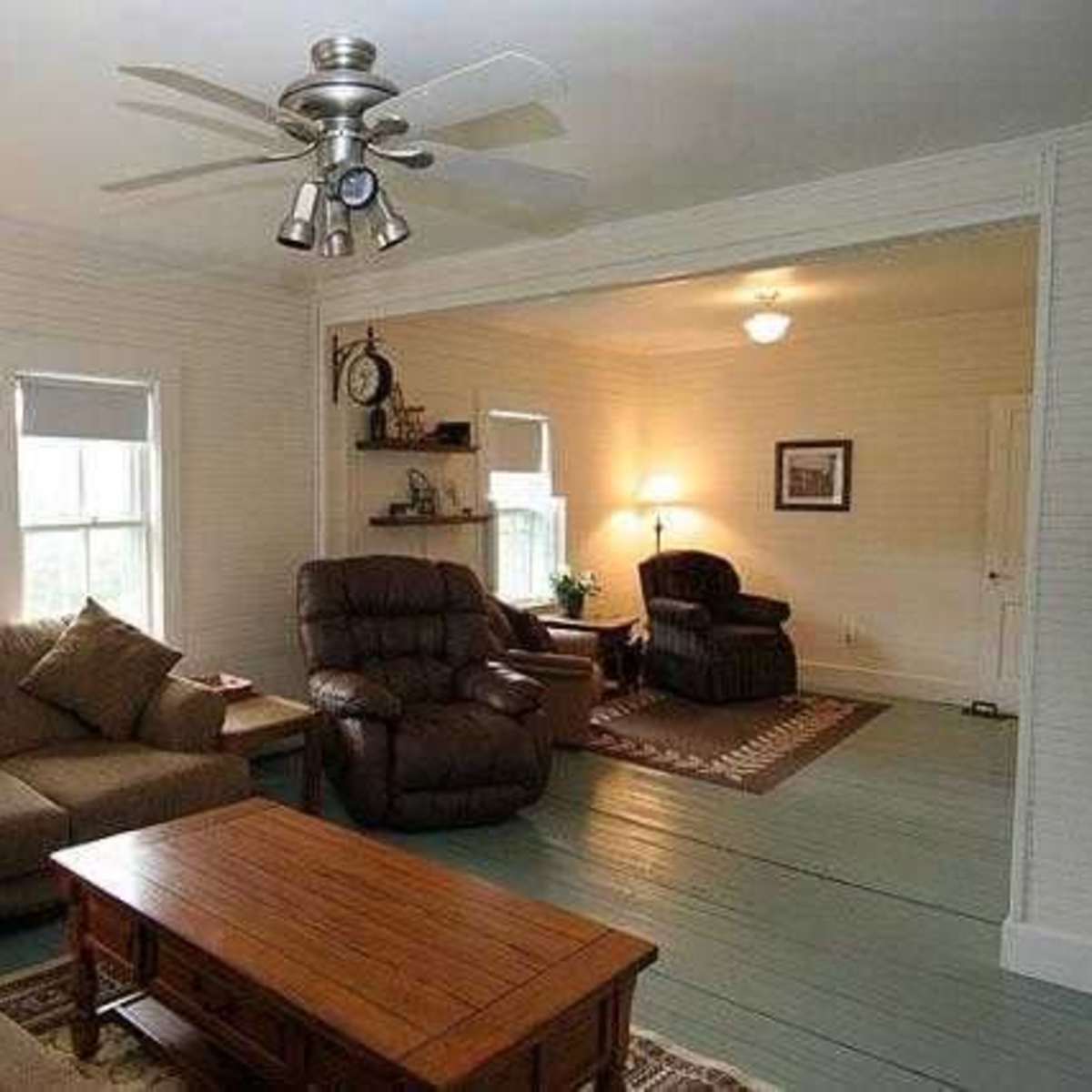 A living room with a couch, recliner chairs, a wooden coffee table, and light-colored walls, connected to another room through an archway.