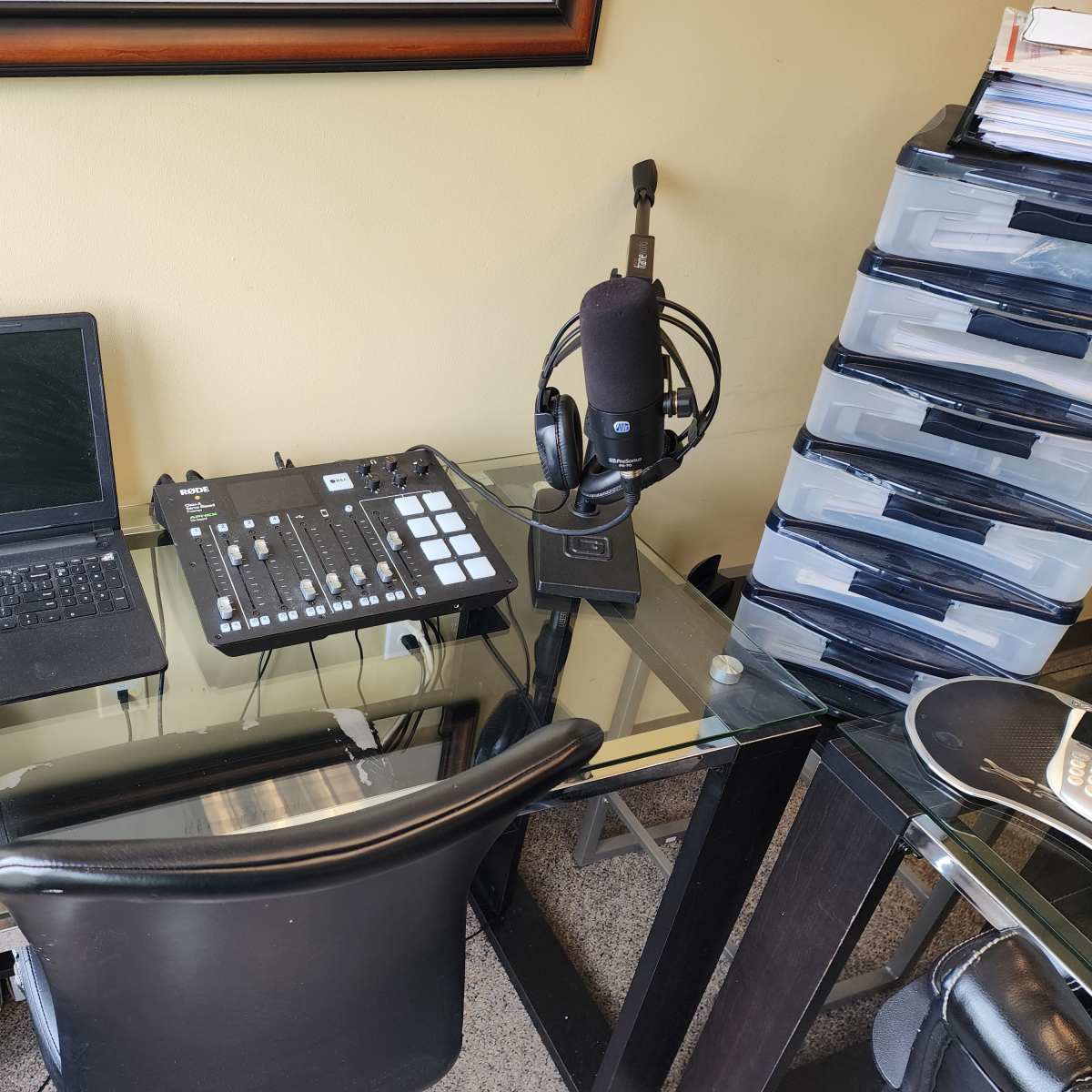 A home office setup features a laptop, audio mixer, microphone, and storage drawers on a glass desk.