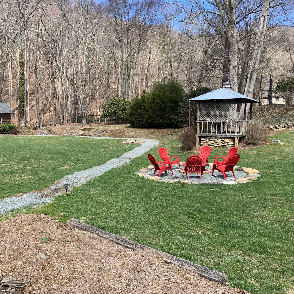 A grassy area with a stone path leading to a circular fire pit surrounded by red chairs, with a cabin in the background and trees beyond.