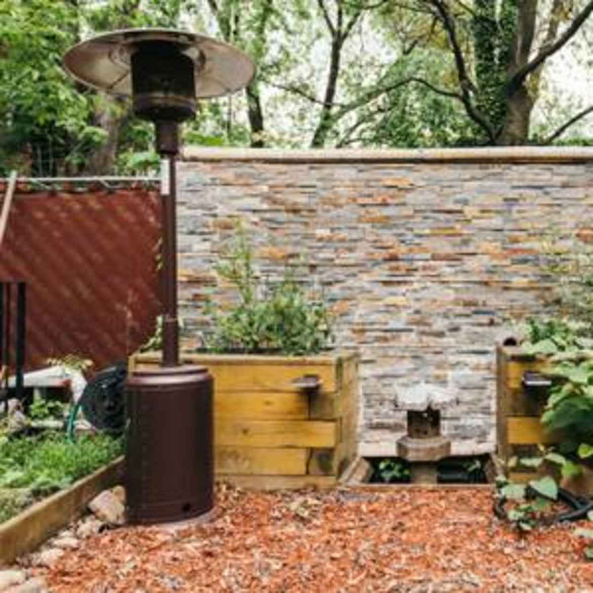 A garden area featuring a stone wall, wooden planters with plants, a gas heater, and a grill.
