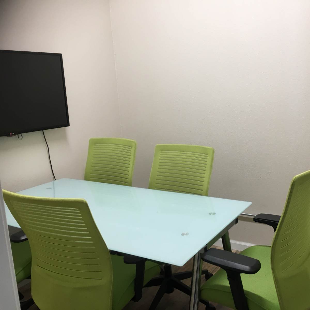 A small meeting room features a glass table surrounded by four green chairs and a wall-mounted television.