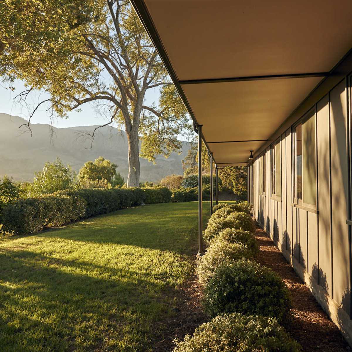 A long building with windows, bordered by well-maintained hedges and a grassy lawn, set against a backdrop of mountains and trees.