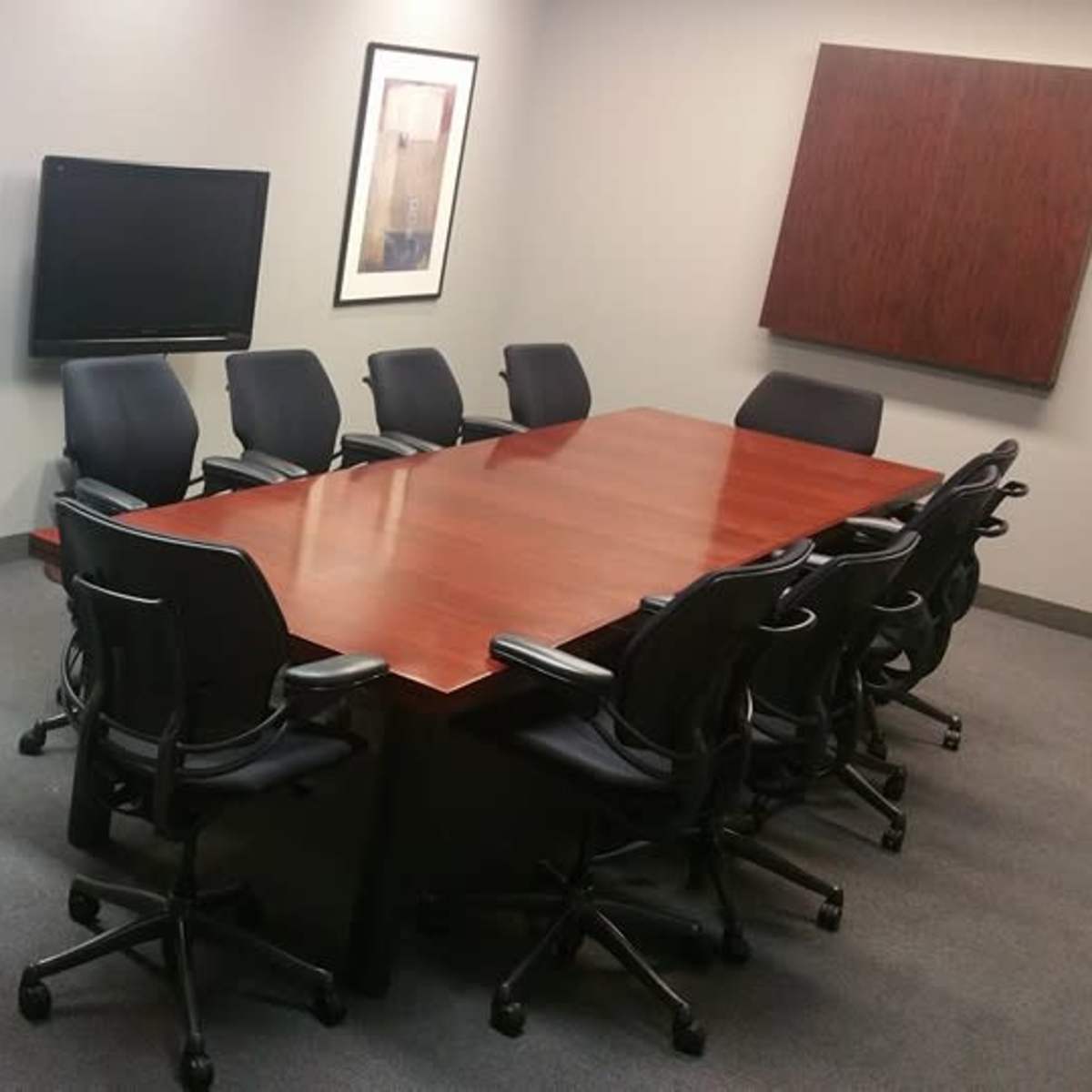 A conference room features a large wooden table surrounded by black rolling chairs, with two framed artworks and a television mounted on the wall.