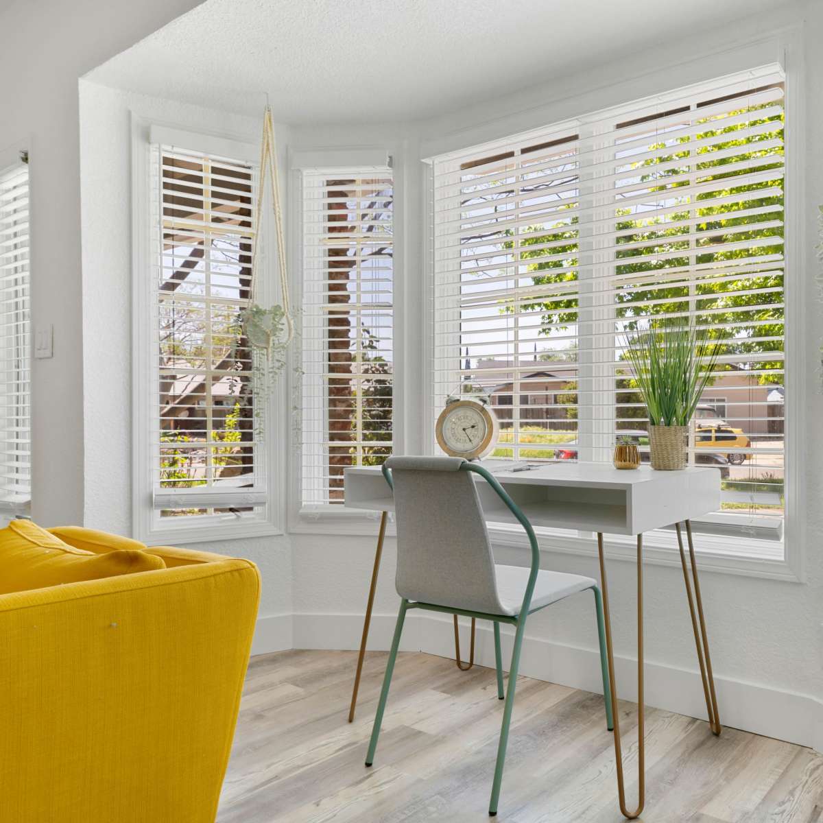 A home office desk with a chair sits by a bay window adorned with blinds, offering a view of a street lined with trees.