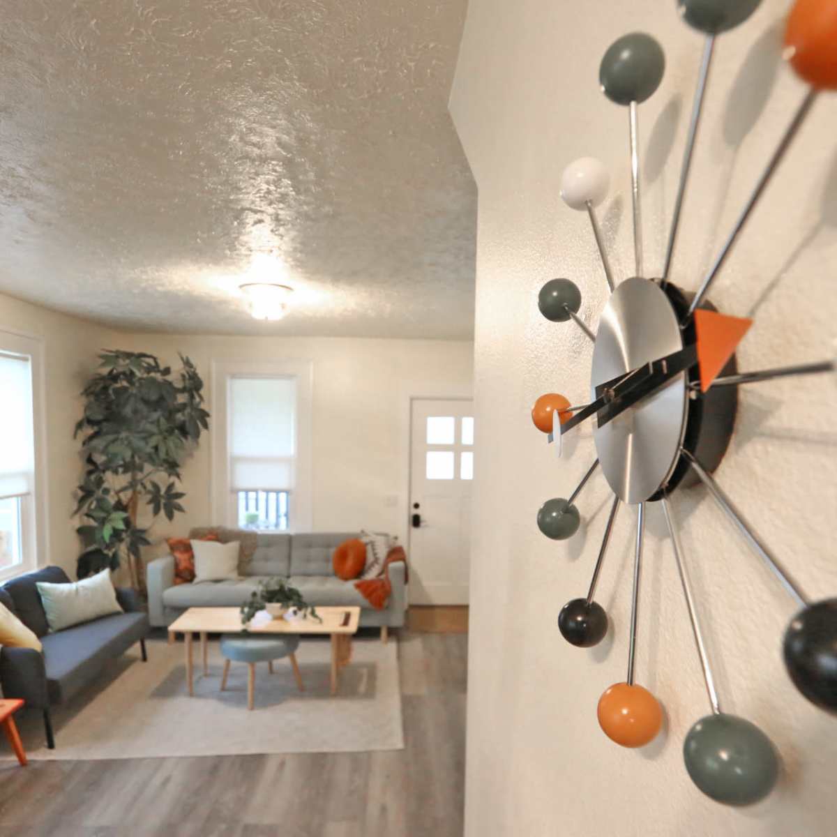 A modern living room features a gray sofa, a coffee table, and a large plant, all seen from an angled perspective that includes a colorful wall clock.