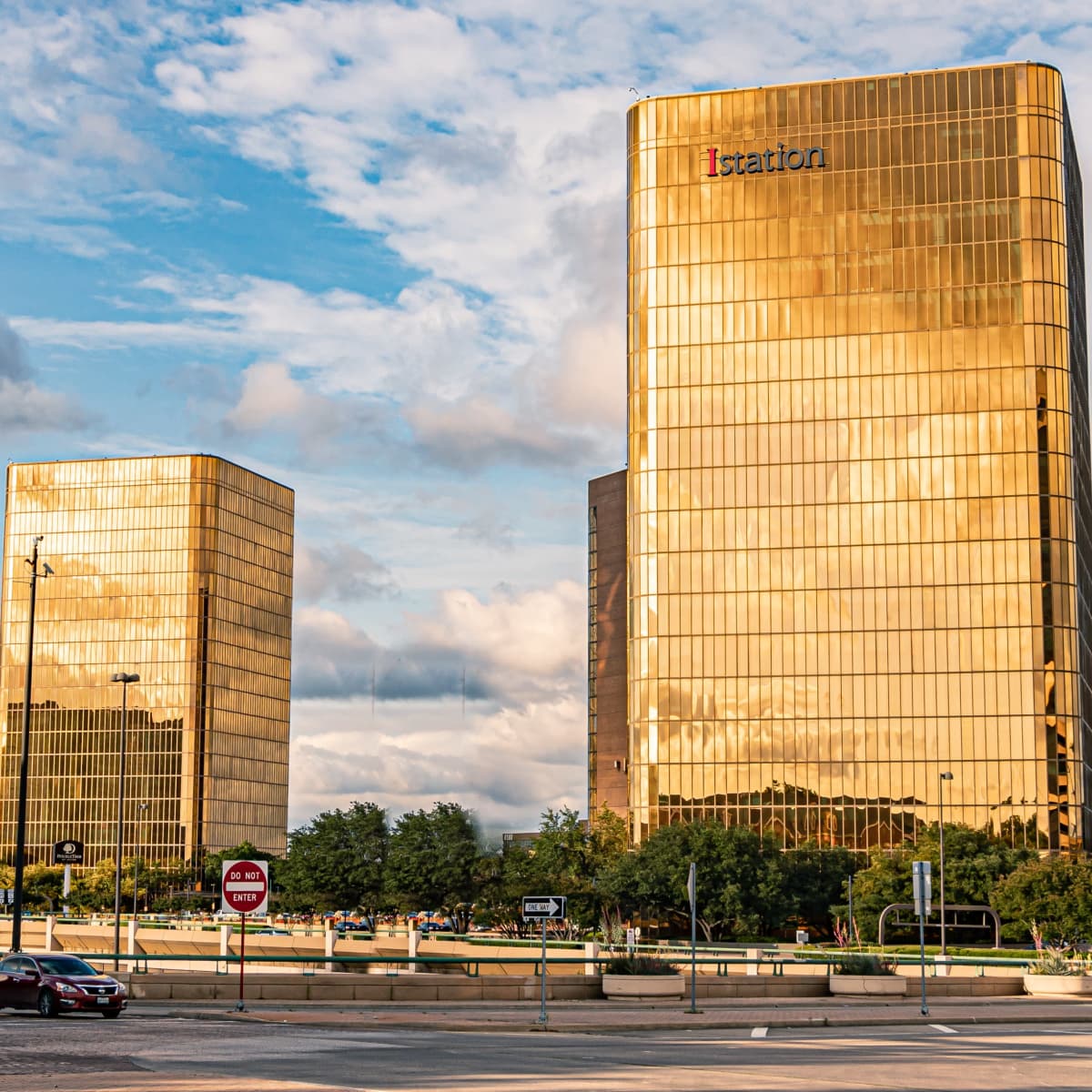 Two tall glass buildings reflect sunlight in a cityscape, with traffic and greenery visible in the foreground.
