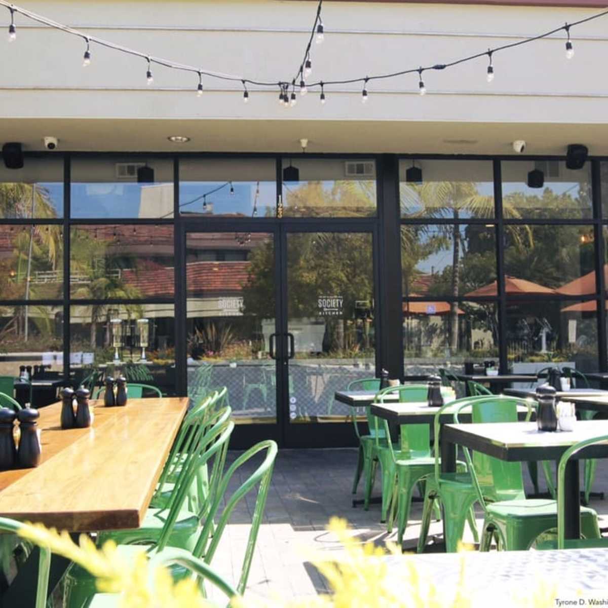 A patio area with green metal chairs and wooden tables, overlooking large glass doors that reflect the surroundings.