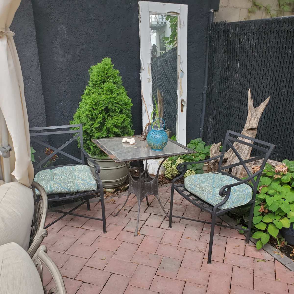 A small outdoor seating area features two metal chairs and a table set on a brick patio, with a mirror and potted plants in the background.
