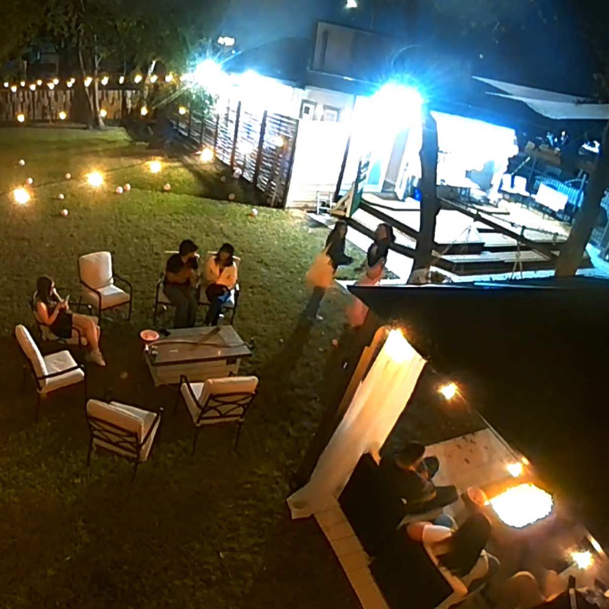 A dimly lit outdoor area with several seating arrangements, string lights overhead, and people gathered around various tables and seating spaces.