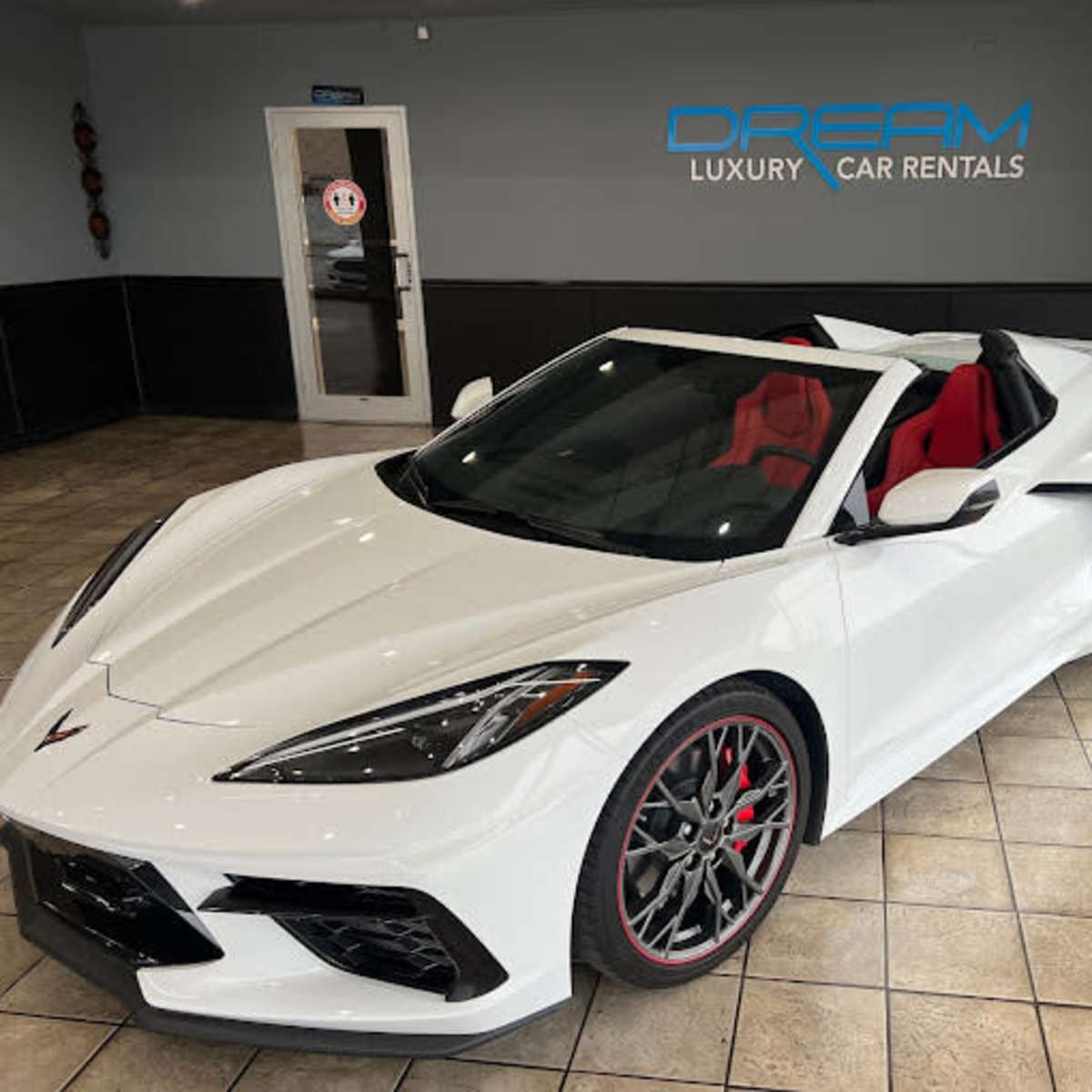 A white convertible sports car with red seating is parked inside a rental facility featuring a sign that reads "DREAM LUXURY CAR RENTALS."