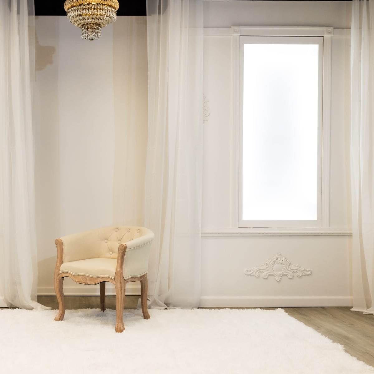 A single upholstered chair is positioned in the center of a brightly lit room with large windows and sheer curtains.