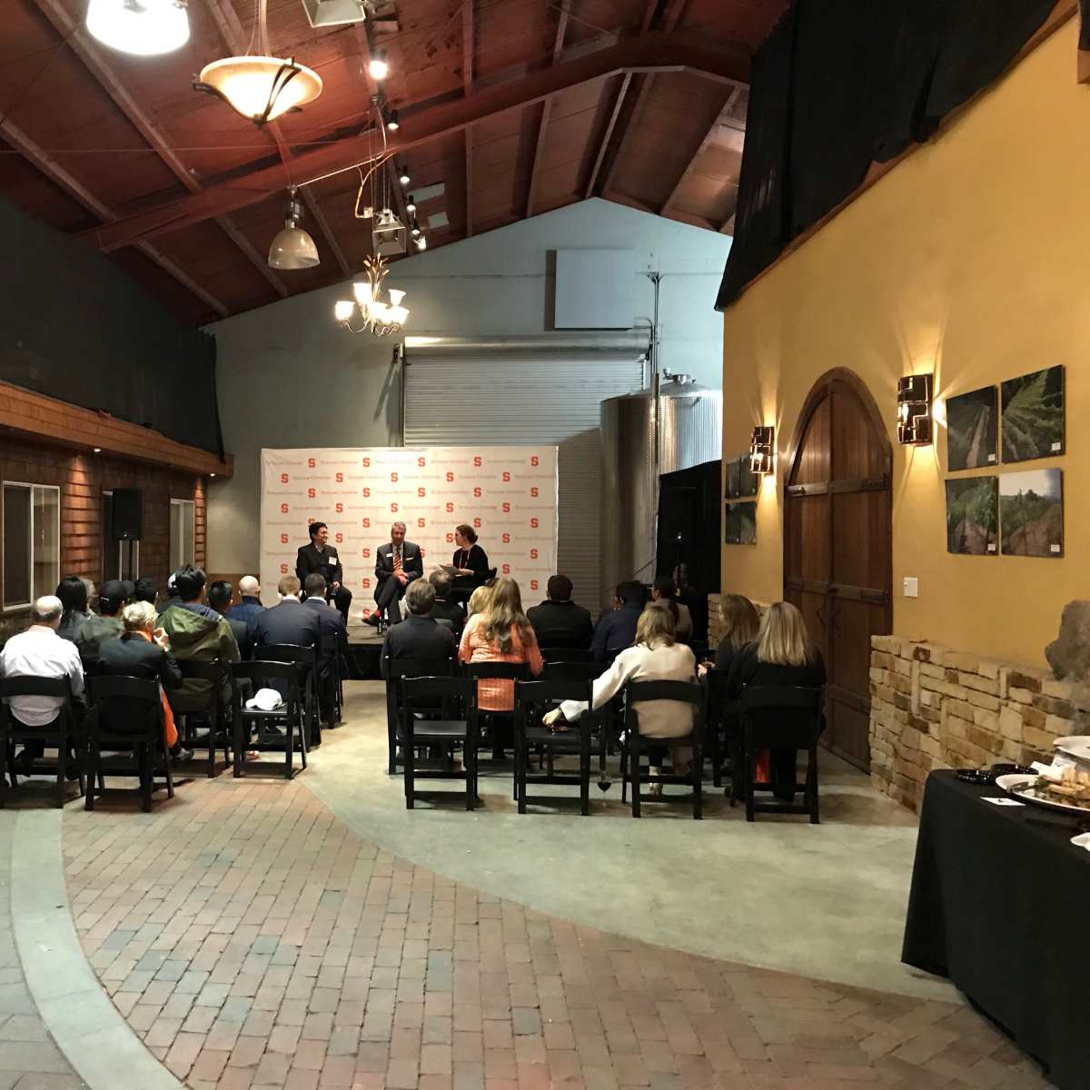A group of people sits in chairs facing a panel at a presentation event indoors, with refreshments on a table to the side.