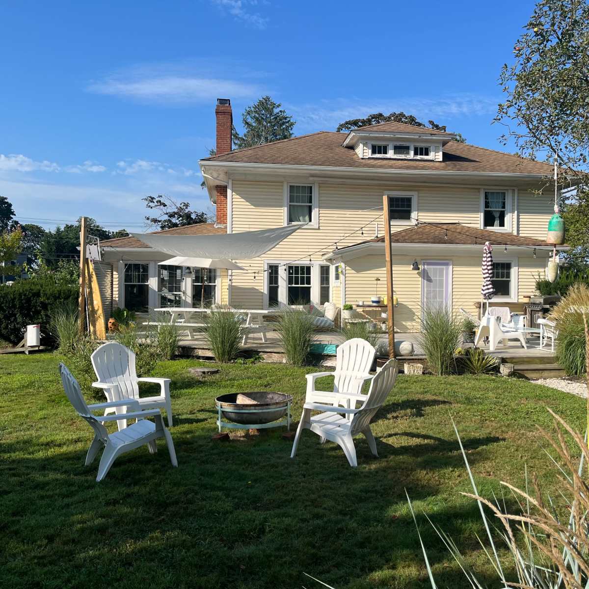 A large yellow house with a broad porch and a fire pit surrounded by white chairs sits in a grassy yard under a clear blue sky.