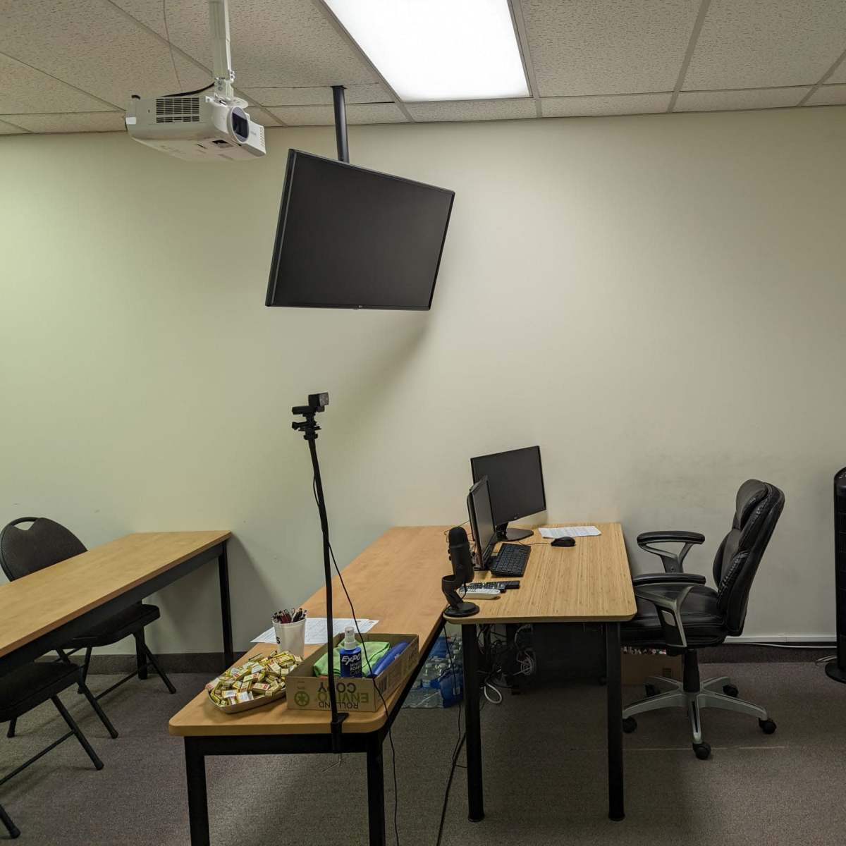 A classroom with two tables, an empty office chair, a computer setup, a projector mounted on the ceiling, and a whiteboard on the wall.