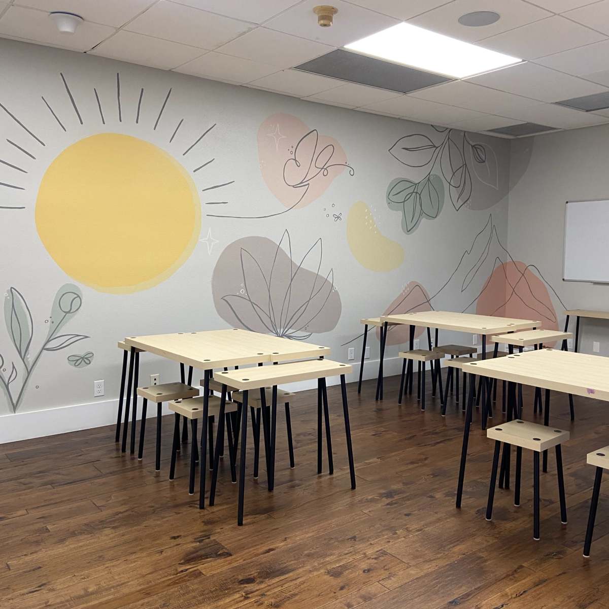 The room features light-colored walls with a mural of a sun and abstract plants, along with several wooden tables and stools arranged for seating.
