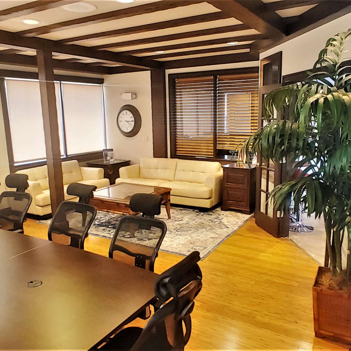 A modern office space featuring a conference table with chairs, a lounge area with a sofa, and large windows with blinds, along with a wall-mounted display screen.