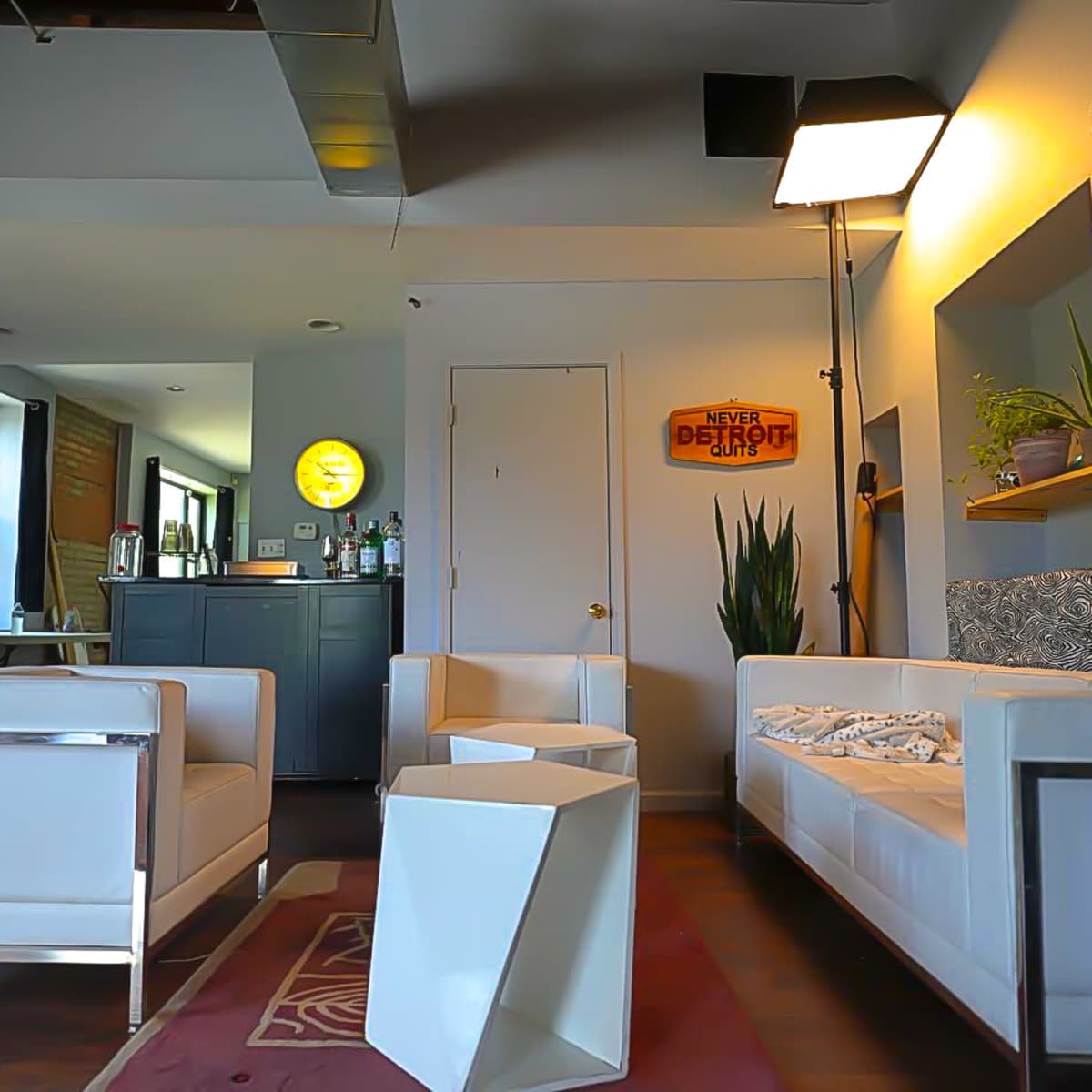 A modern lounge area featuring two white sofas, a geometric white side table, a bar counter with liquor bottles, and framed artwork on the walls.