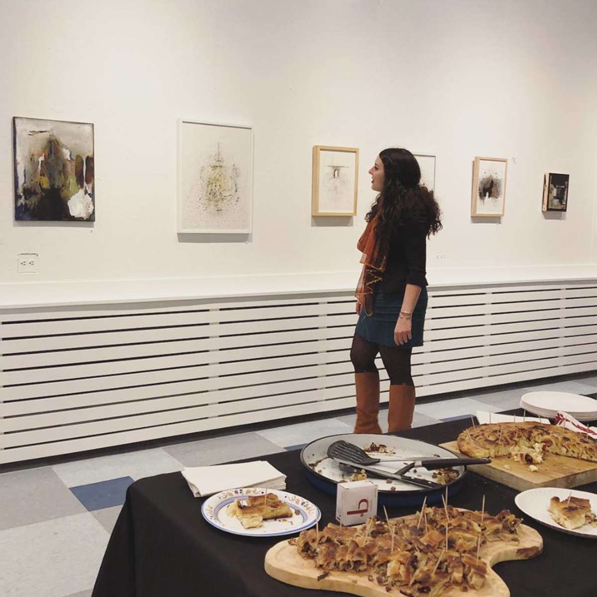 A woman stands in an art gallery, examining the framed artworks on the wall while a table with food is set in the foreground.