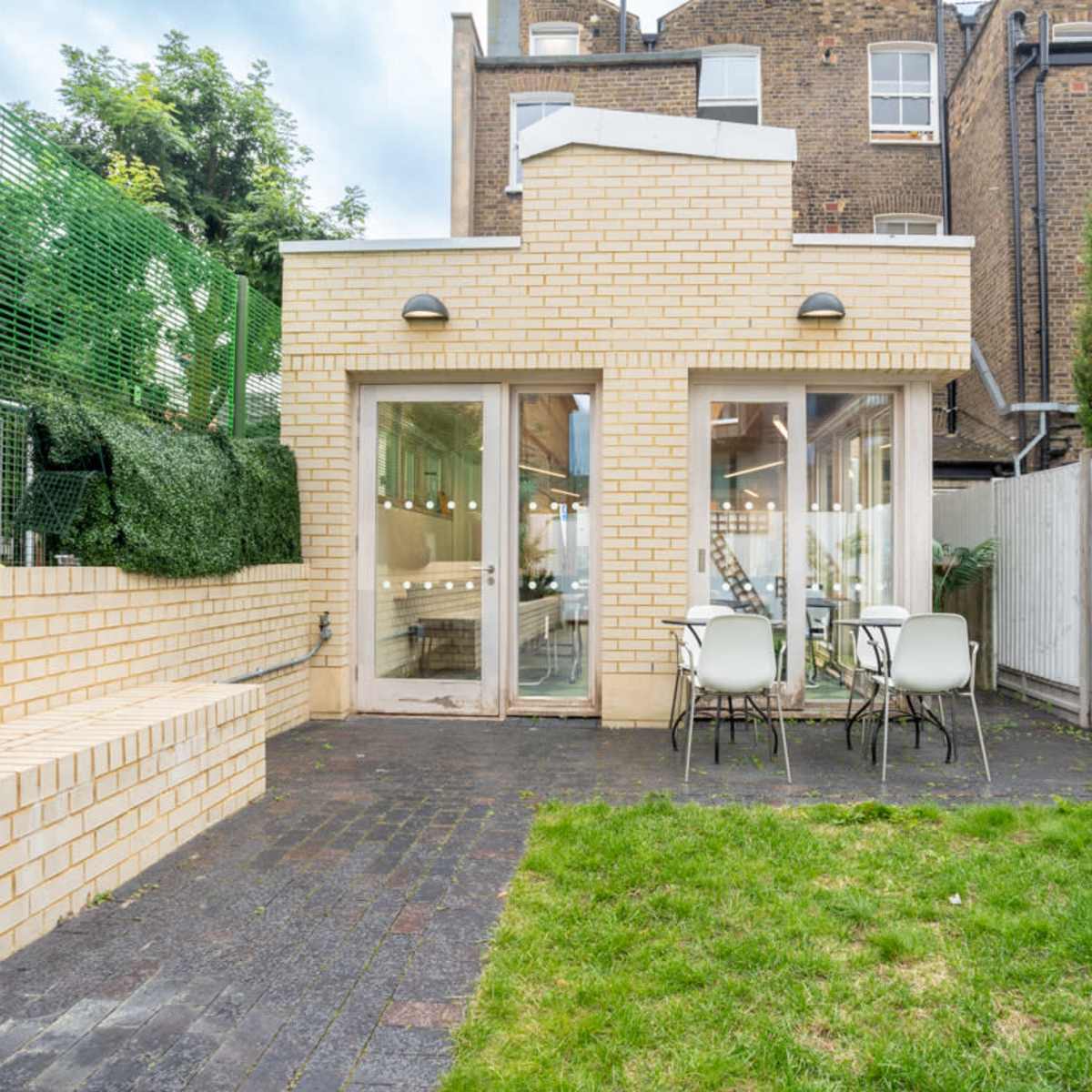 A modern brick extension features large glass doors opening to a small outdoor seating area with two chairs and a grassy space.