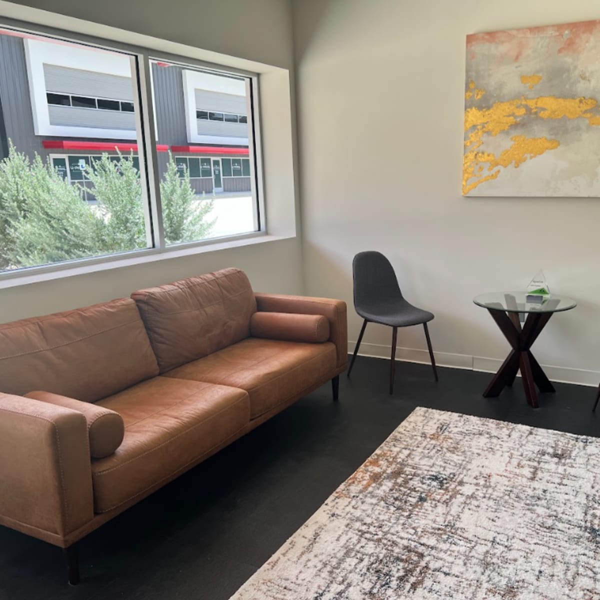 A modern waiting area featuring a brown leather sofa, two chairs, a small glass table, and an abstract painting on the wall.