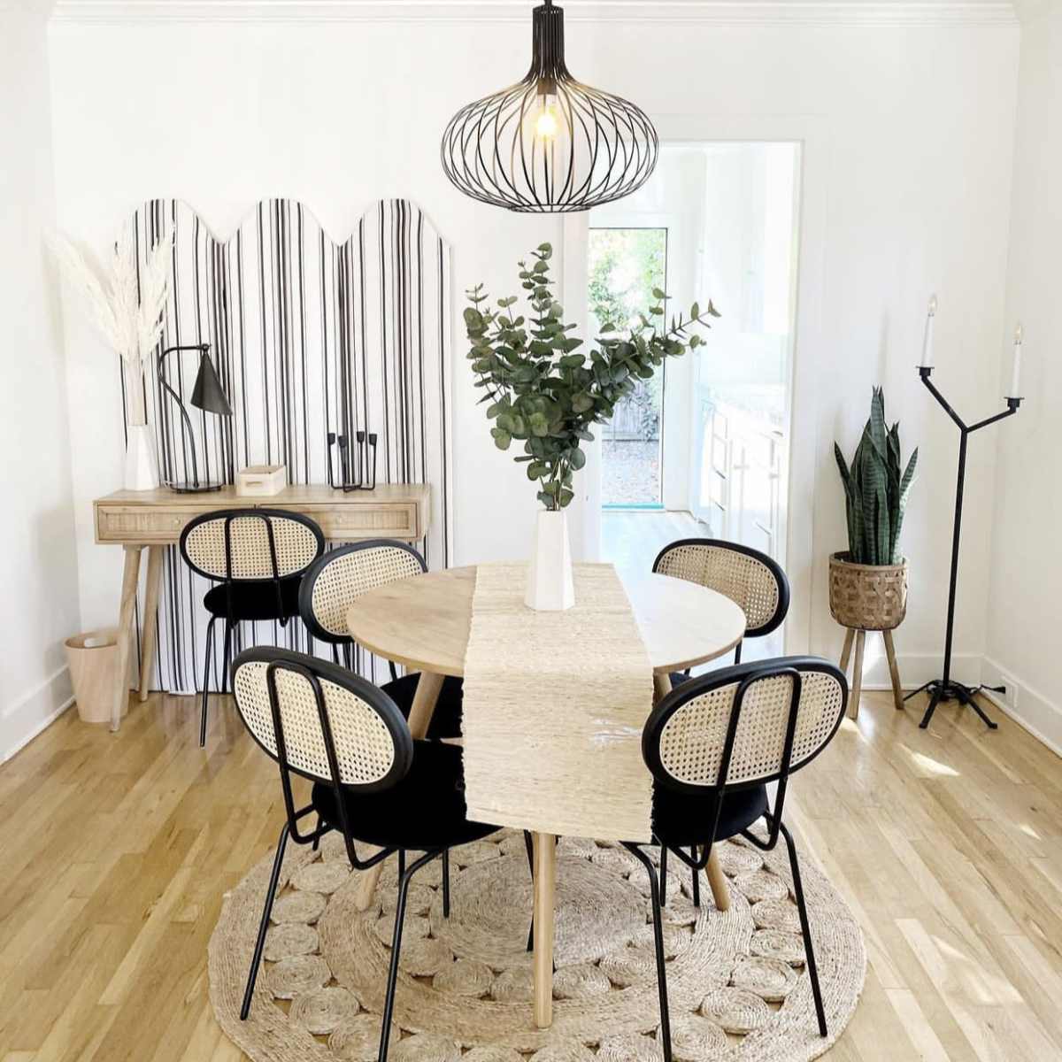 A modern dining area features a round table surrounded by six black and rattan chairs, with a decorative vase of greenery on the table and a patterned area rug beneath it.