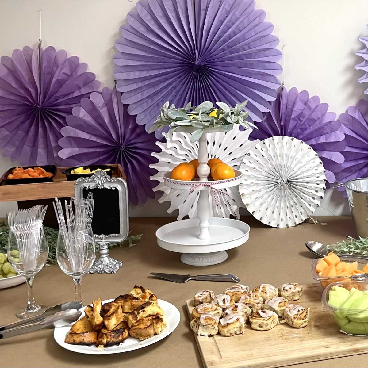 A festive table setup with a variety of food items, including baked goods and fresh fruit, alongside decorative paper fans on the wall.