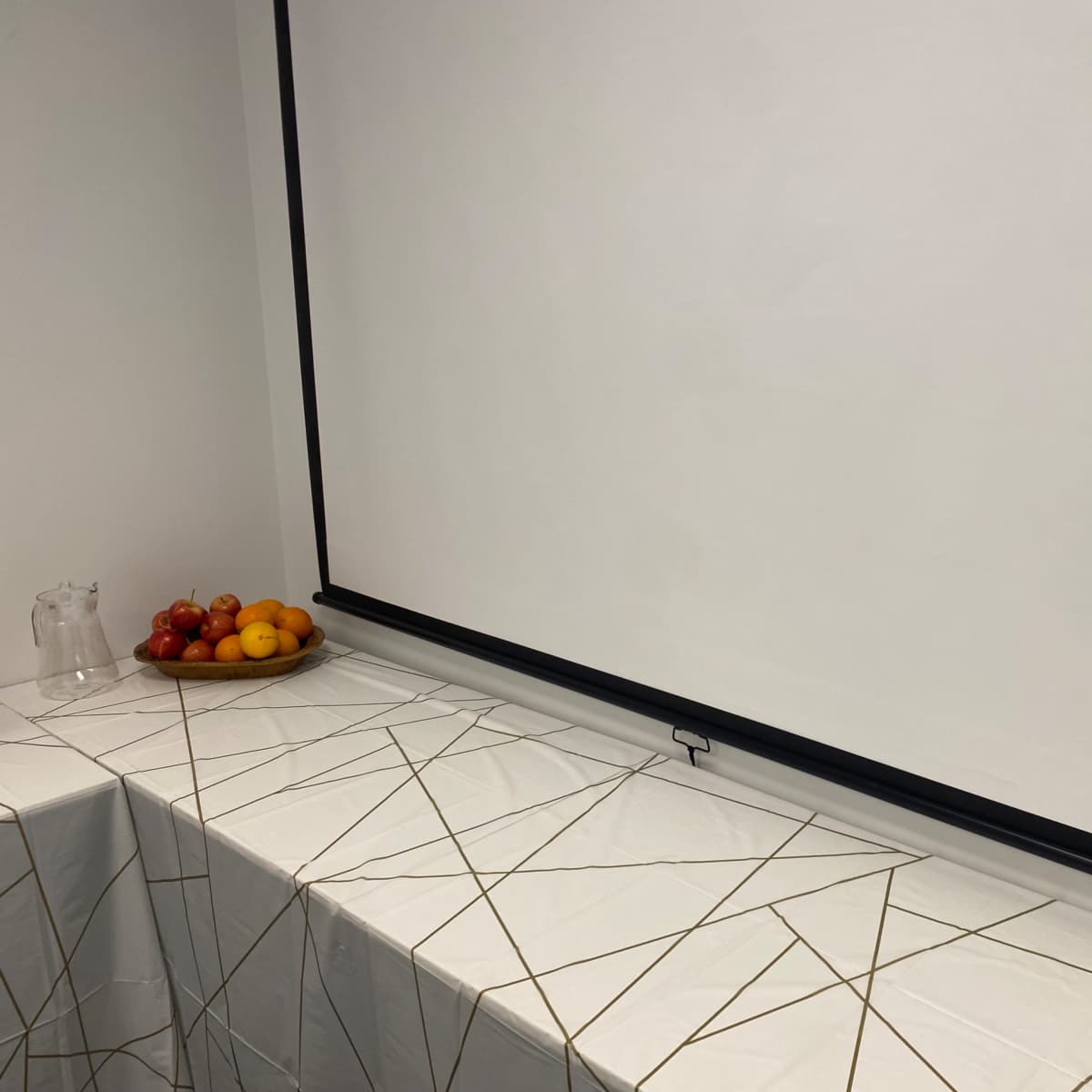 A table covered with a white tablecloth featuring a geometric pattern holds a bowl of fruits next to a blank wall-mounted projection screen.