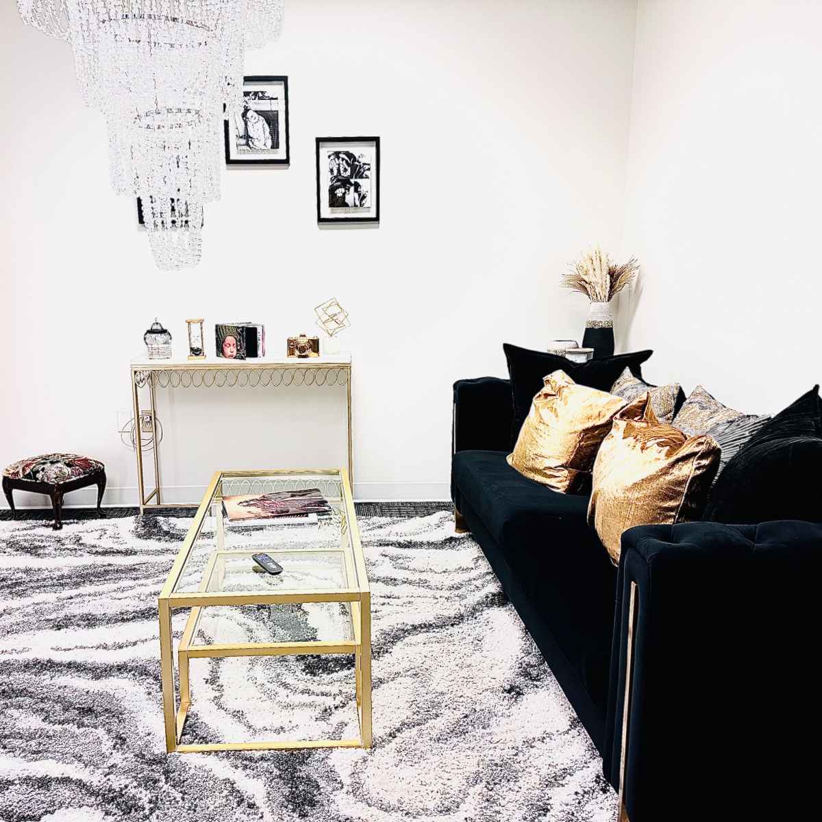 A stylish interior featuring a black sofa with decorative cushions, a gold-framed glass coffee table, a dress form, and a chandelier, all set on a patterned rug.