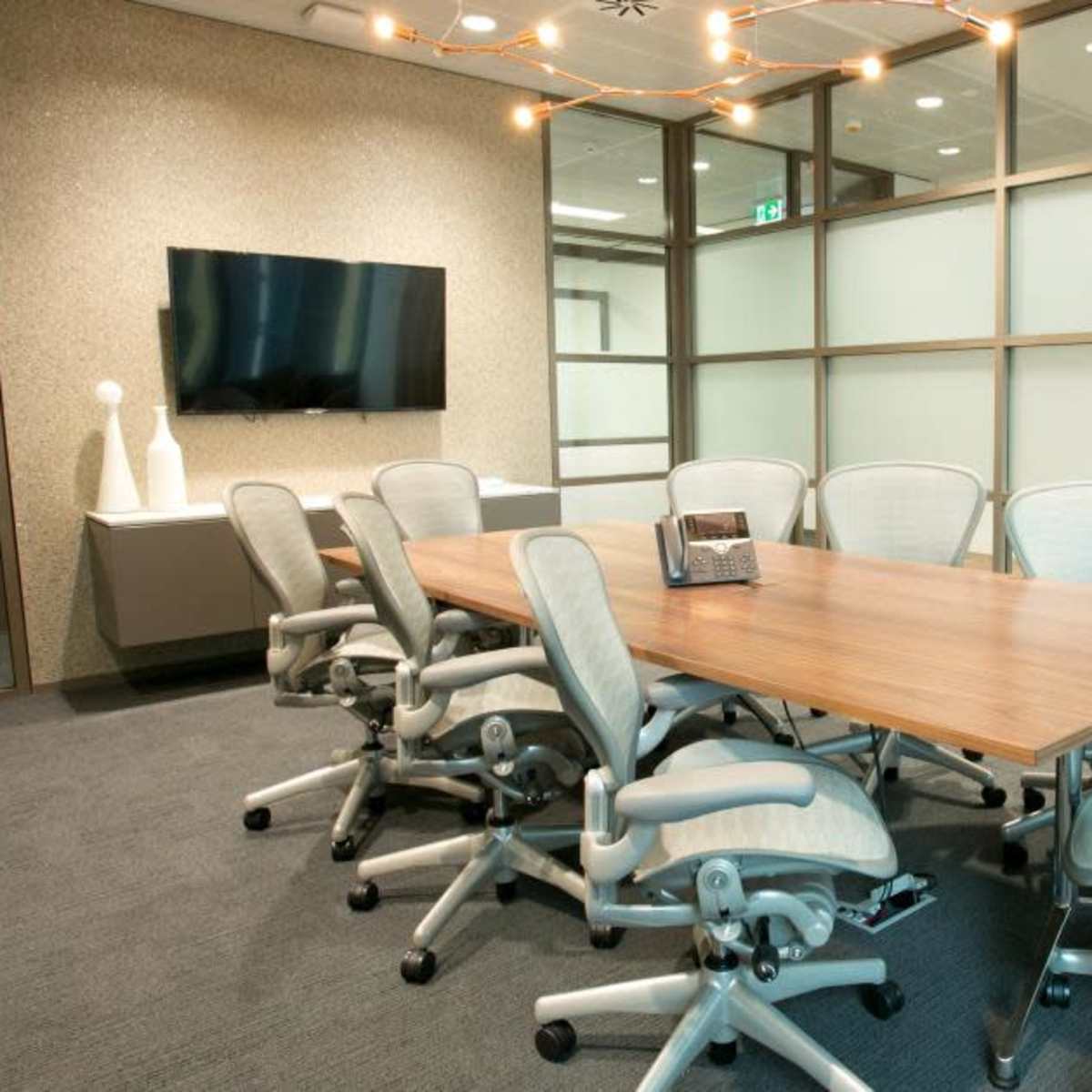 A conference room features a wooden table surrounded by ergonomic chairs, a wall-mounted screen, and a decorative light fixture.