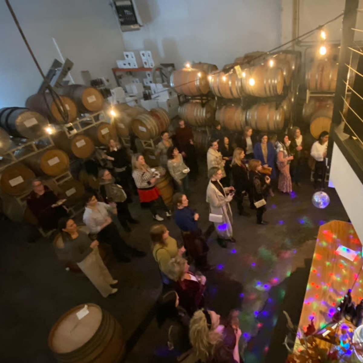 A crowd gathers in a dimly lit winery, surrounded by wooden barrels, while a colorful light display reflects off a disco ball.