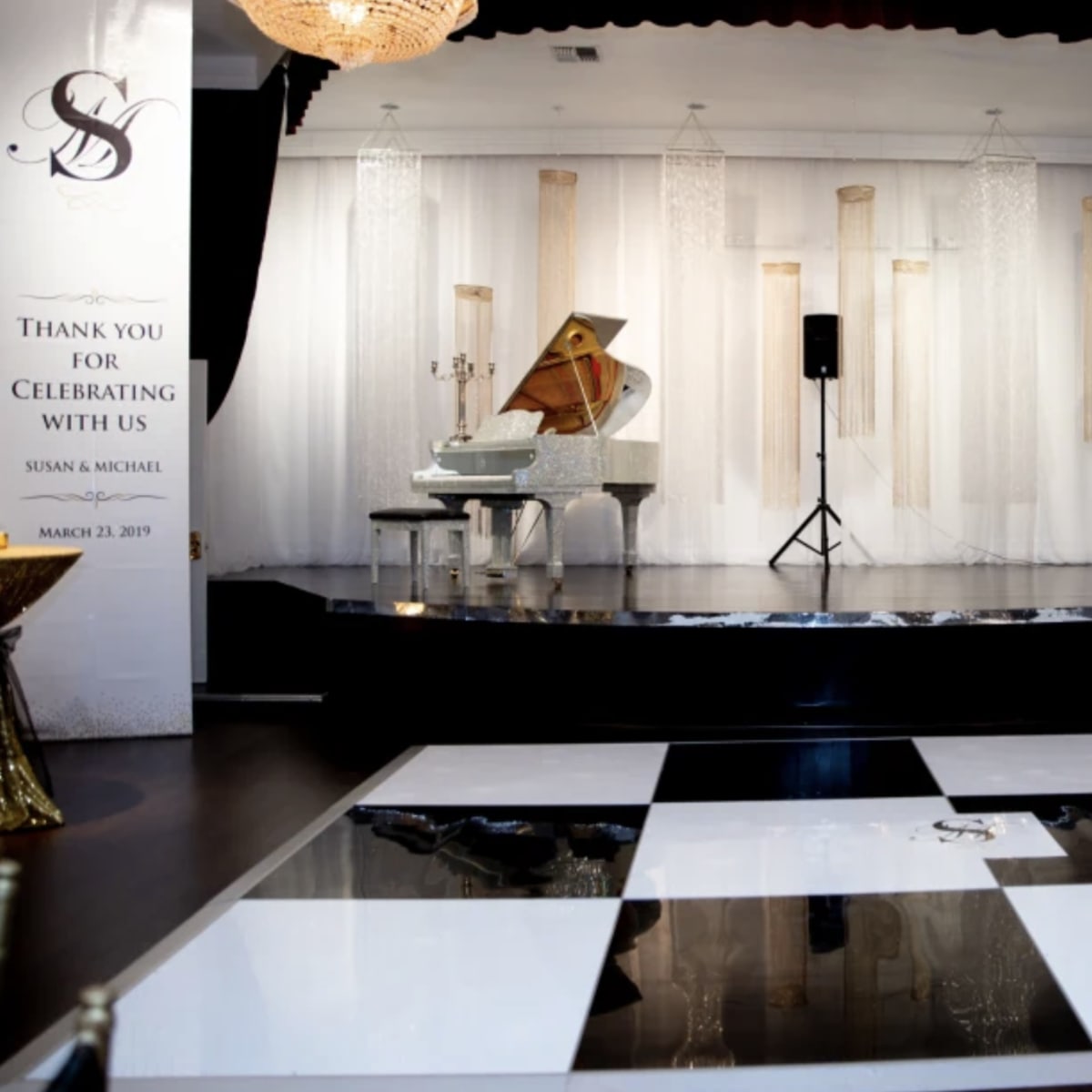 A decorated event stage with a grand piano, speakers, a black and white dance floor, and elegant drapery in the background.