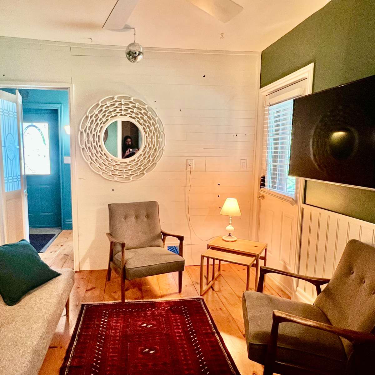 A cozy living room with a gray sofa and two armchairs facing a wooden side table and a wall-mounted television, while a mirror and a door lead to another room.