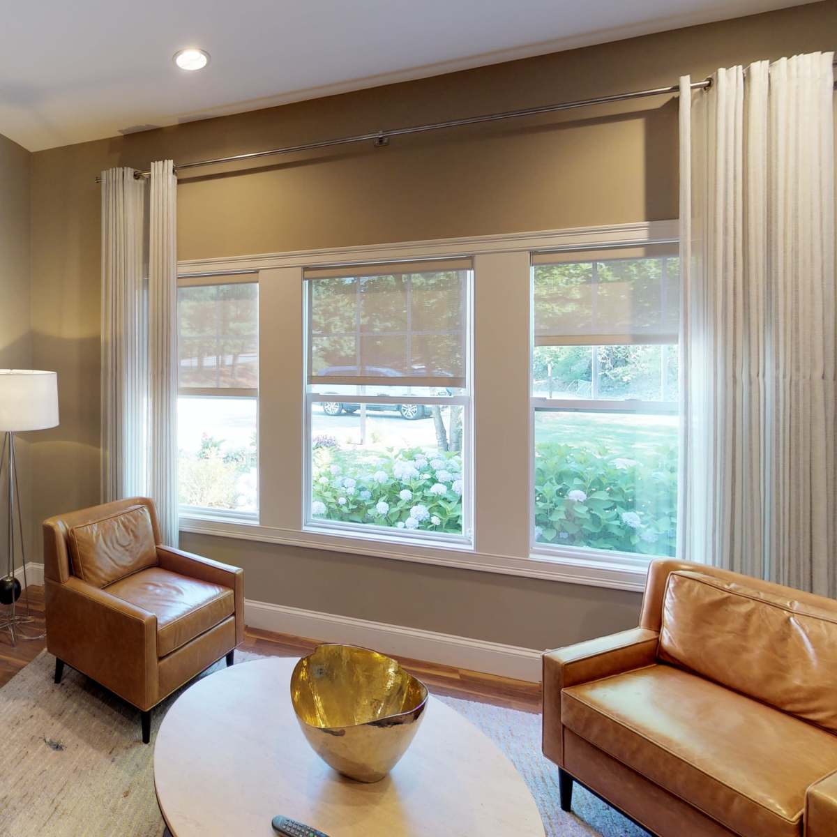 A living room with two brown leather chairs, a round table, a lamp, and large windows that allow natural light to enter.