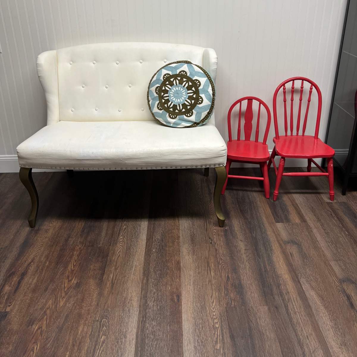 A white settee with a decorative pillow is positioned next to two red wooden chairs in a room with wooden flooring.
